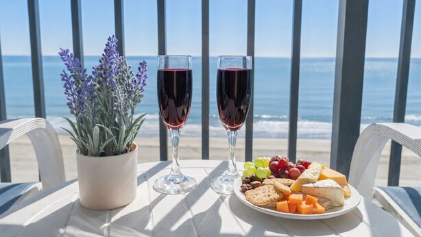 A table with two wine glasses, a potted plant, and a plate of cheese and grapes on a balcony overlooking the sea.