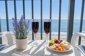 Ein Tisch mit zwei Weingläsern, einem Blumentopf und einer Käse- und Weintraubenteller auf einem Balkon mit Blick aufs Meer.