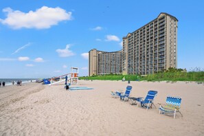 A beach with a large hotel in the background, a lifeguard tower, and several beach chairs.
