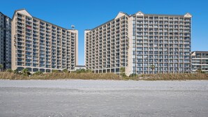 A beachfront hotel with multiple high-rise buildings, featuring balconies and a sandy beach in the foreground.