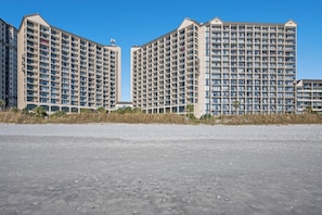Un hôtel en bord de mer composé de plusieurs immeubles de grande hauteur, doté de balcons et offrant une vue sur une plage de sable au premier plan.