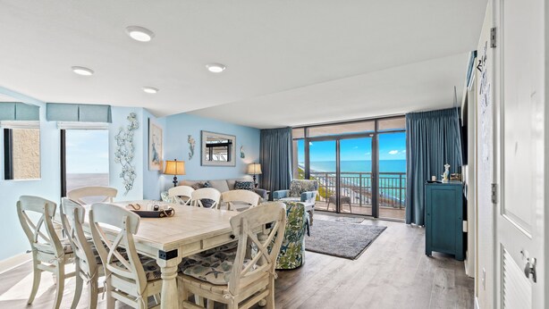 A dining area with a wooden table and chairs, a view of the ocean through large windows, and a balcony with a view.
