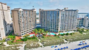 A hotel with multiple high-rise buildings, a beach area with umbrellas, and a pool.