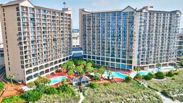 A hotel with multiple high-rise buildings, a swimming pool, and a landscaped area with palm trees. - Beach Cove Resort 1202 - 2 Bed/2Bath Ocean Front Condo (North Myrtle Beach)