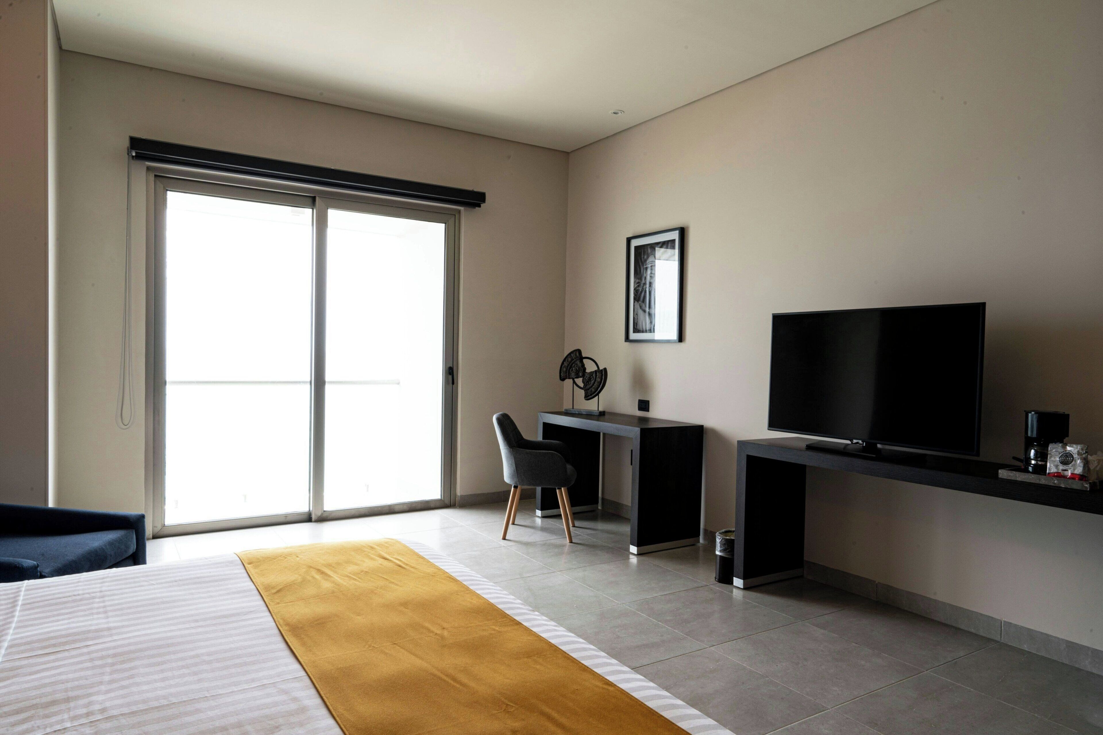 Una habitación de hotel moderna con una cama, un escritorio, una silla, un televisor y un ventanal grande.