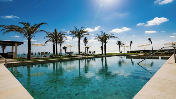 A clear swimming pool with lounge chairs and umbrellas, surrounded by palm trees and a view of the ocean. - Delta Hotels by Marriott Riviera Veracruz (Alvarado)