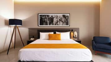 Quarto de hotel com uma cama, mesa de cabeceira, luminária, cadeira e um quadro pendurado na parede.