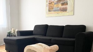 A living room with a black sectional sofa, a beige ottoman, and a painting on the wall. - Grand T2 Lumineux Pied du Métro et Centre de Lyon (Villeurbanne)