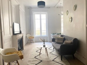 Un salon avec un canapé, une table basse et un téléviseur. On y trouve également une cheminée, une fenêtre et une porte.