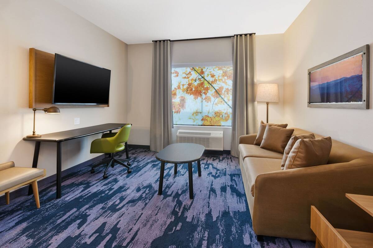 Ein Hotelzimmer mit Flachbildfernseher, einer Couch, einem Schreibtisch mit Stuhl, einem kleinen Tisch und einem Fenster mit Blick auf Herbstlaub.