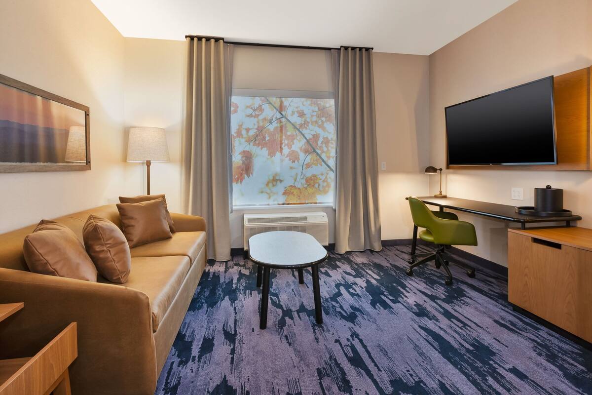 Ein Hotelzimmer mit einer Sitzecke, einem kleinen Tisch, einem Schreibtisch, einem Fernseher und einem Fenster mit Vorhängen.