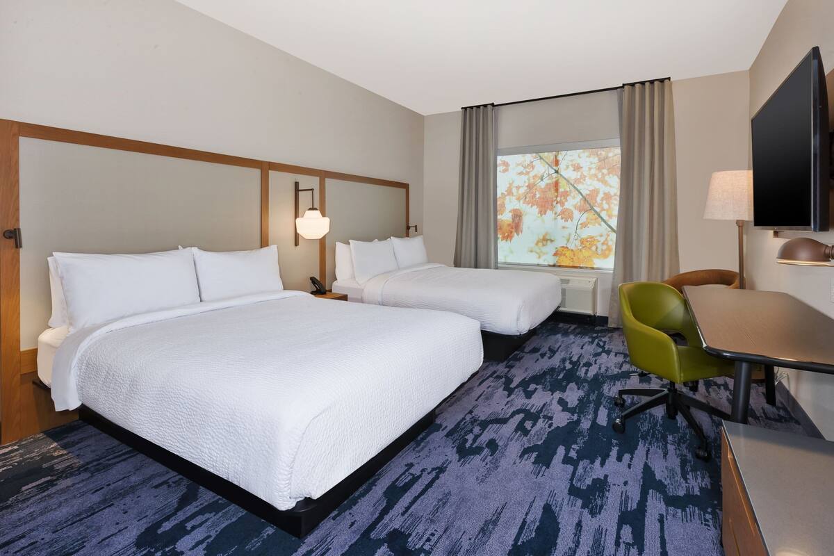 Ein Hotelzimmer mit zwei Betten, einem Schreibtisch, einem Sessel, einem Fernseher und einem Fenster mit Vorhängen.
