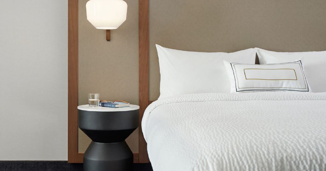 A hotel room with a bed, a bedside table with a lamp, and a nightstand with a glass of water and books.