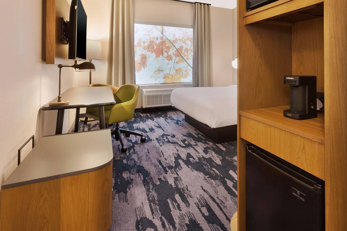 Ein Hotelzimmer mit Bett, Schreibtisch mit Lampe, Stuhl, Kaffeemaschine und Blick auf Herbstlaub durch das Fenster.