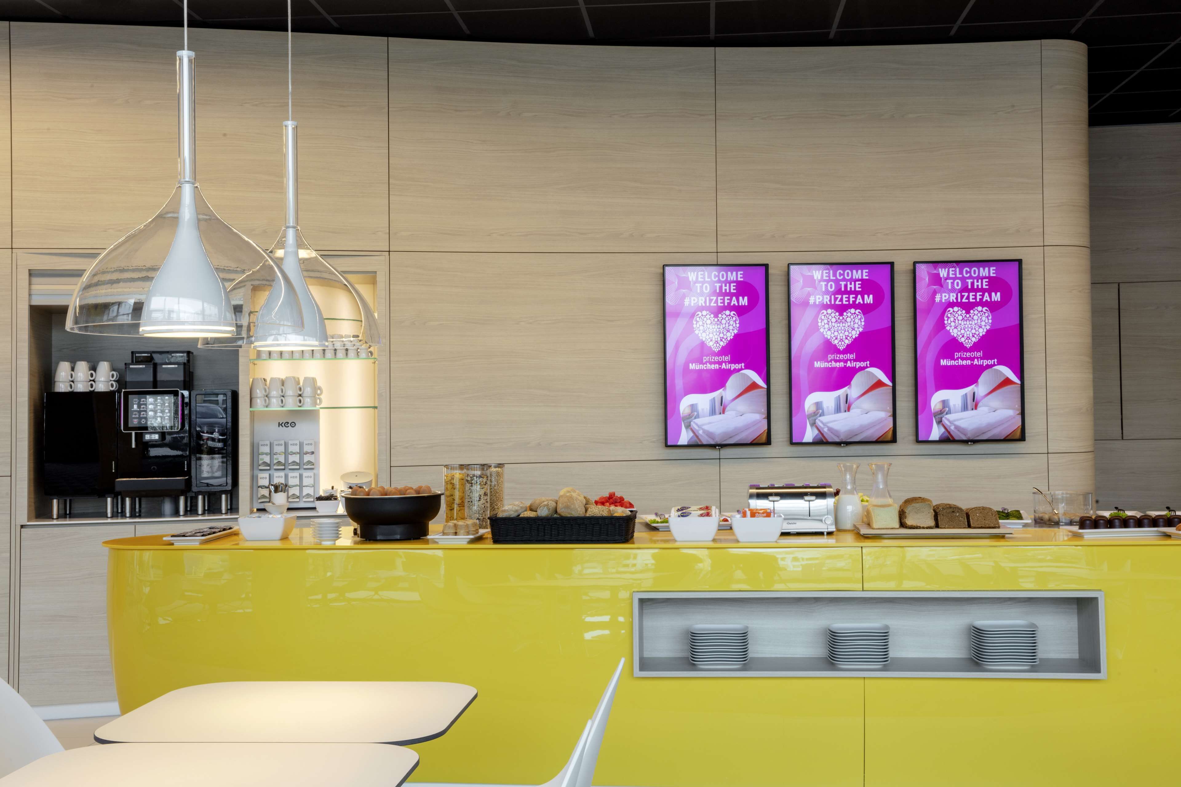 A bright yellow reception desk with a coffee station, three monitors displaying 'Welcome to the #PrizeFam' messages, and a modern interior design.
