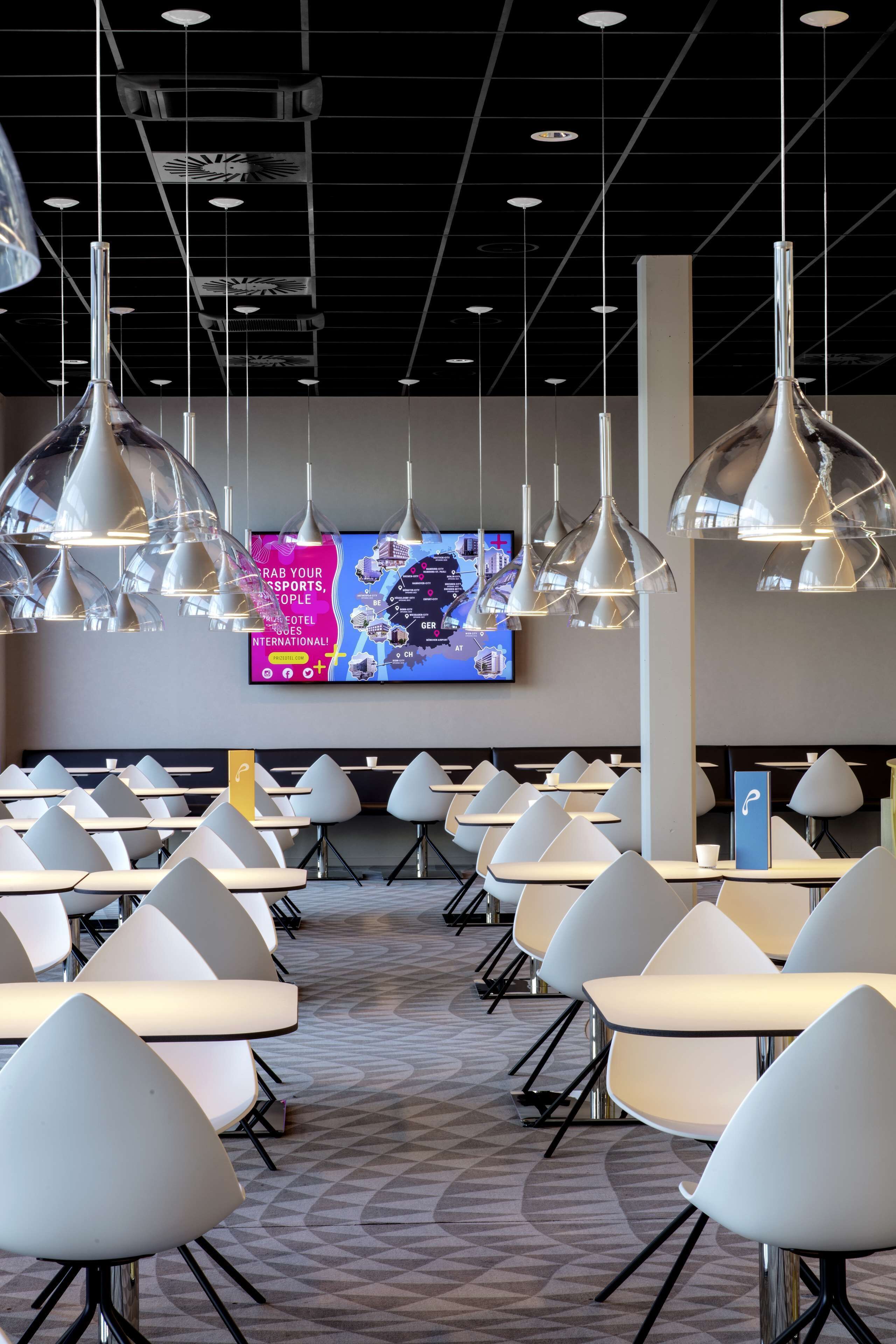 A modern hotel lobby with multiple tables and chairs, a large digital screen displaying a map, and contemporary pendant lighting.