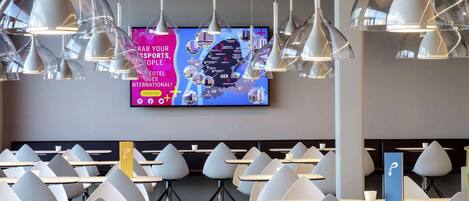 A modern hotel lobby with multiple tables and chairs, a large digital screen displaying a map, and contemporary pendant lighting.