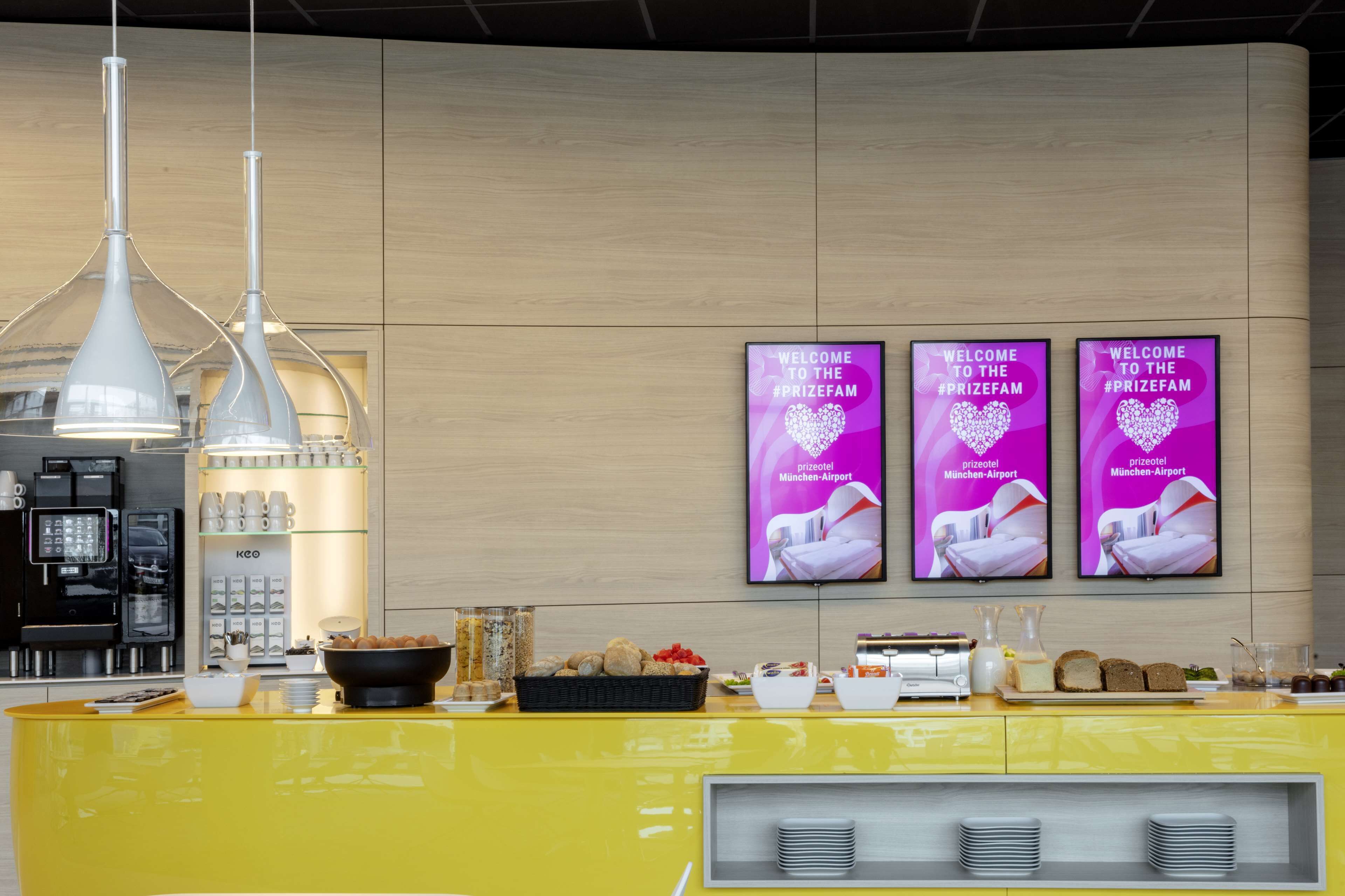 A reception area with a yellow counter, coffee machines, and a wall-mounted TV displaying a welcome message.