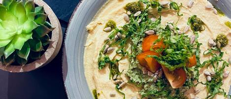 Un tazón de hummus decorado con verduras y frutos secos, una pequeña planta en maceta y una servilleta de tela oscura.