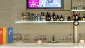 A bar area with a television displaying a map, shelves with liquor bottles, and two black bar stools. - Prize by Radisson, Munich Airport (Hallbergmoos)