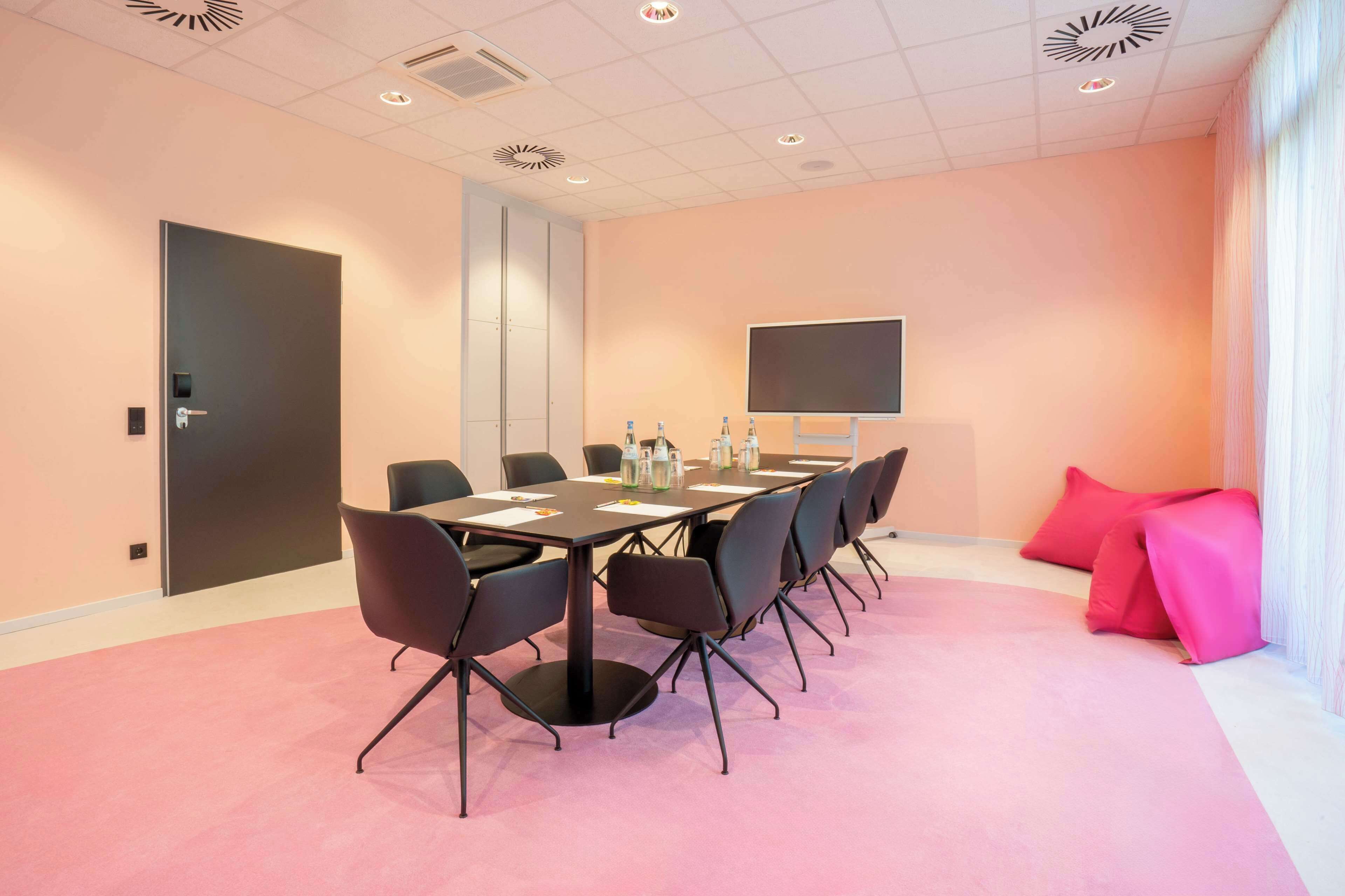 Ett konferensrum med ett långt bord, svarta stolar, en stor skärm och en dörr.