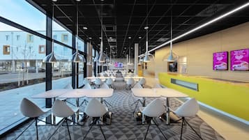 A modern indoor dining area with tables and chairs, large windows, and a yellow accent wall.