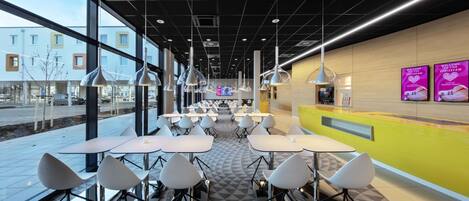 A modern indoor dining area with tables and chairs, large windows, and a yellow accent wall.