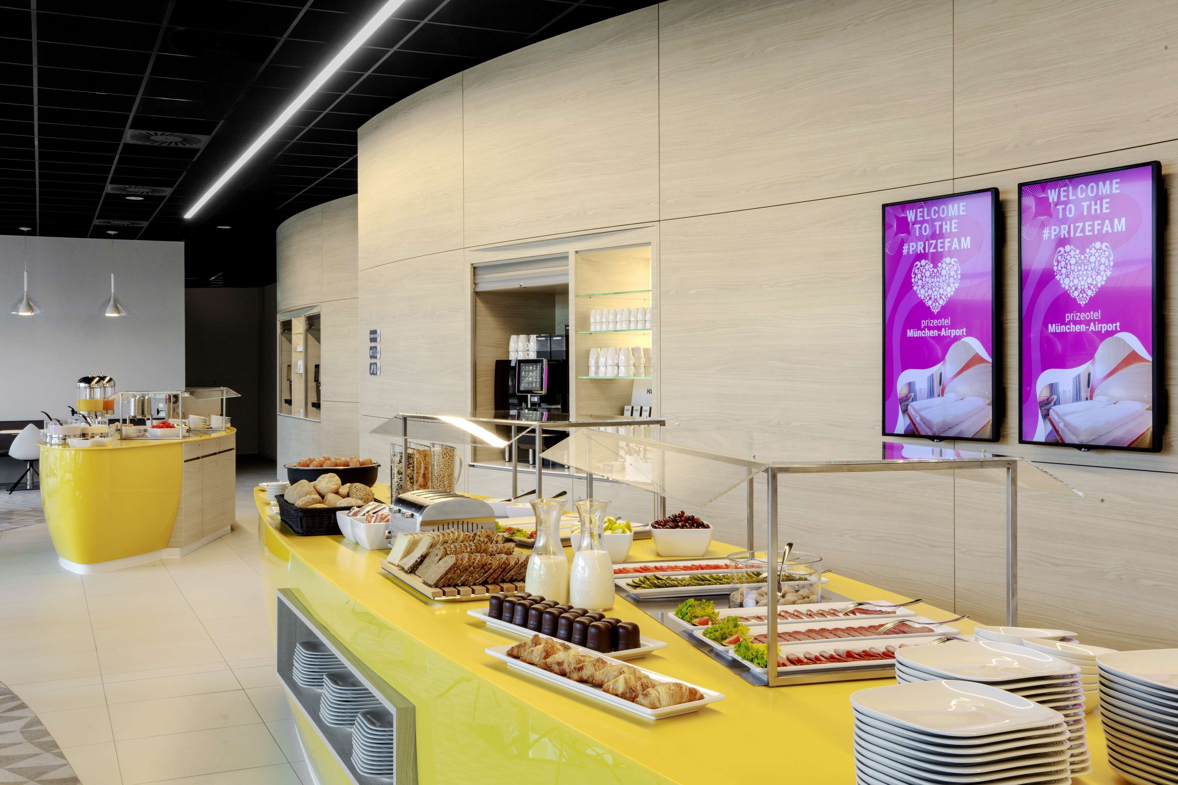 Ein Bereich mit Buffet, verschiedenen Speisen und einer Kaffeestation, gestaltet in modernem Stil mit einem gelben Tresen und Wandmonitoren, die Willkommensbotschaften zeigen.