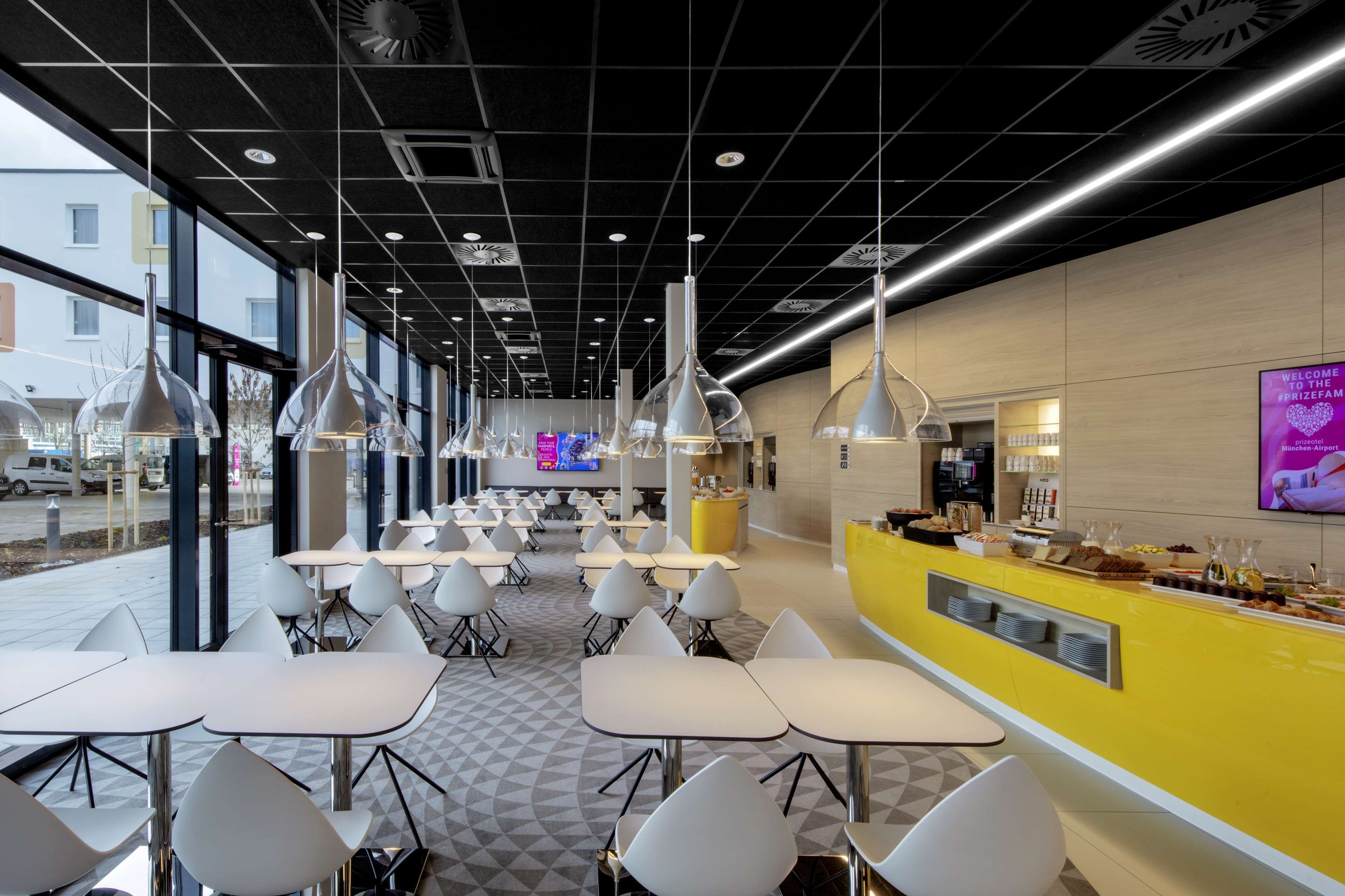 Un café moderno con mesas y sillas blancas, una barra amarilla y una gran pantalla de televisión al fondo.