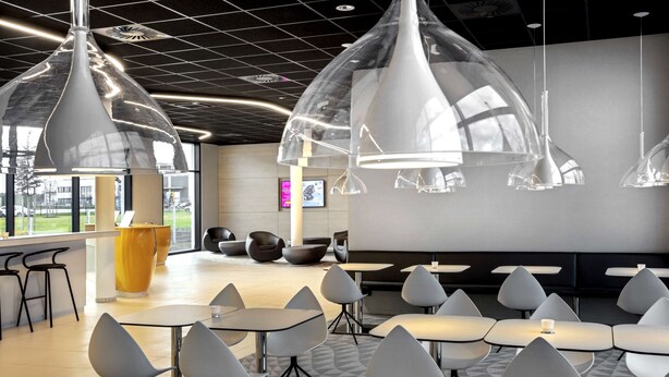 A modern interior with unique pendant lighting, a central yellow vase, and a seating area with tables and chairs. - Prize by Radisson, Munich Airport (Hallbergmoos)