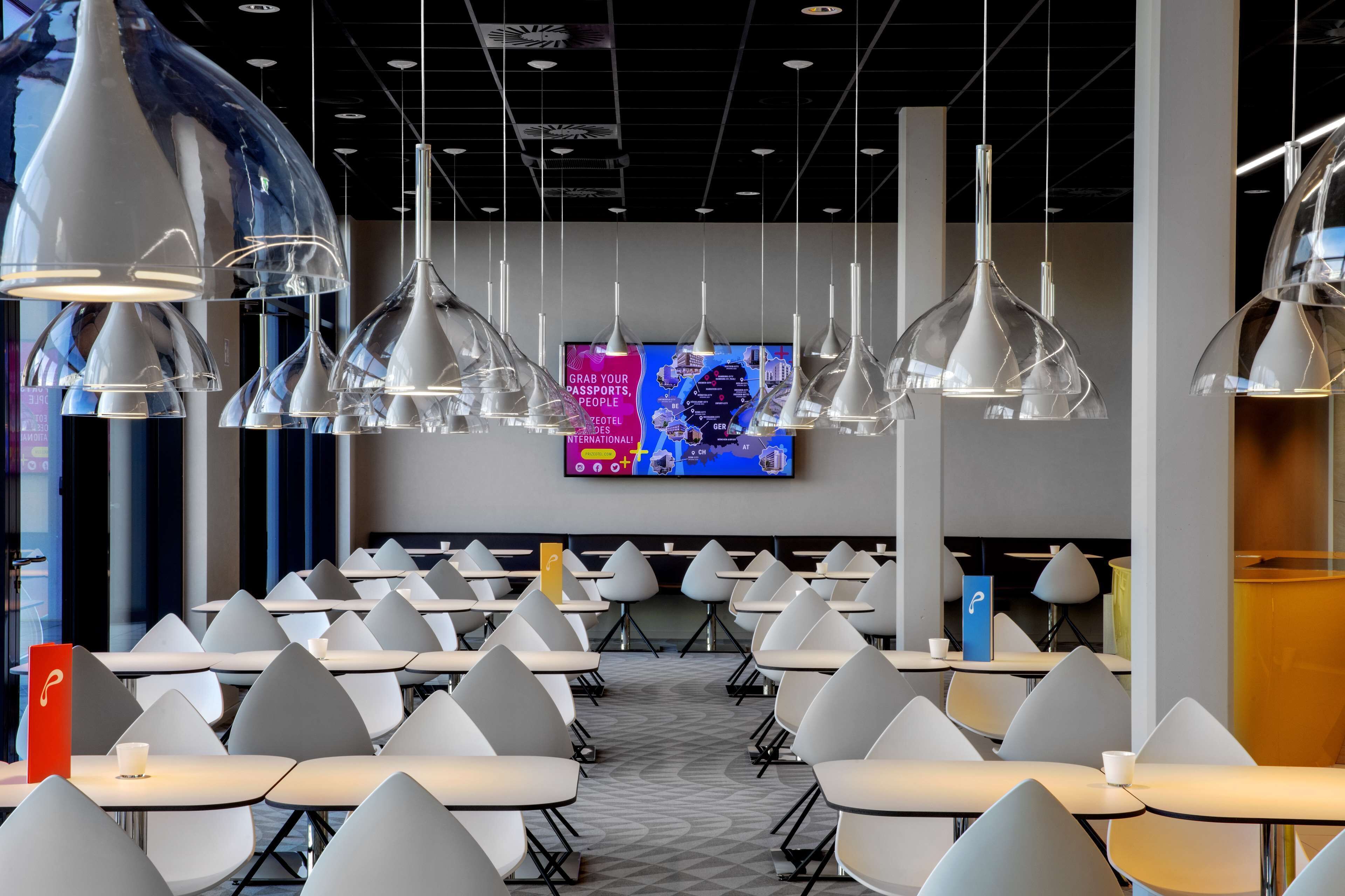 A modern dining area with white tables and chairs, hanging pendant lights, and a large screen displaying a map.