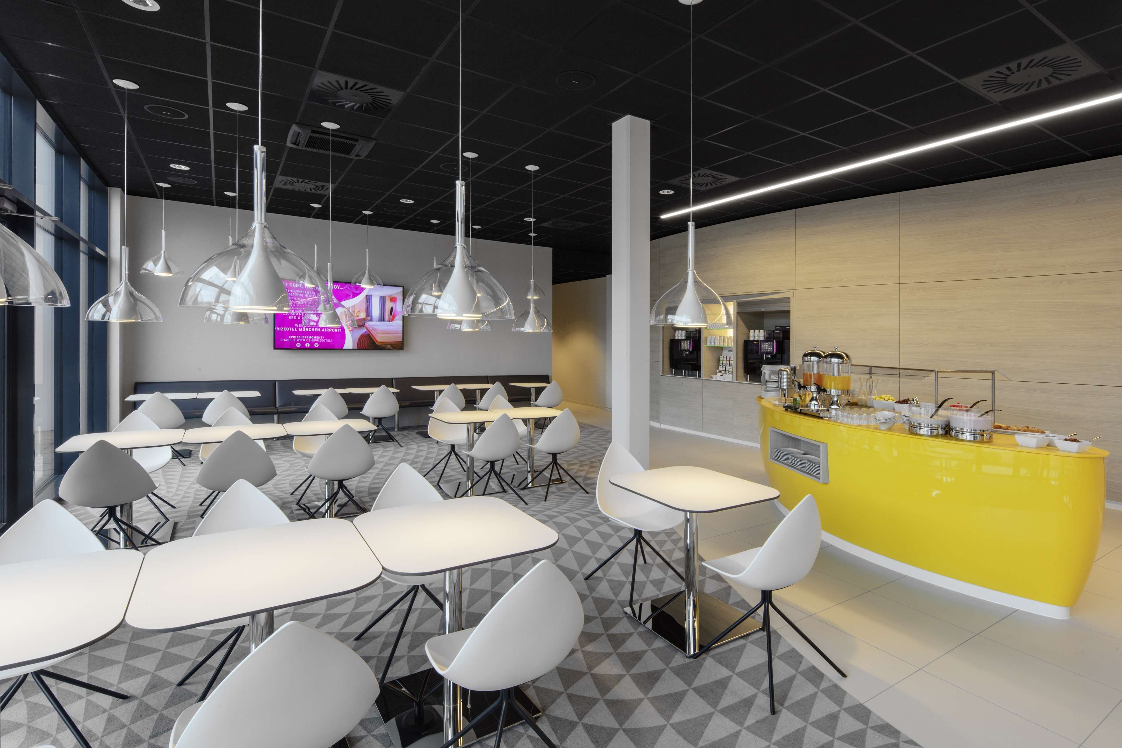 A modern cafeteria with white tables and chairs, a yellow counter with food items, and pendant lighting.