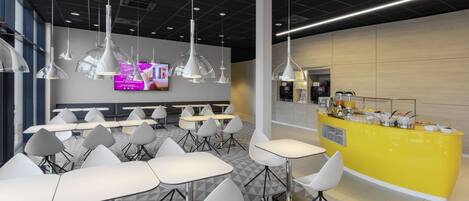 A modern cafeteria with white tables and chairs, a yellow counter with food items, and pendant lighting.