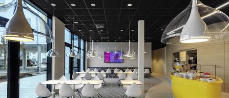 A modern dining area with white tables and chairs, a yellow counter with beverages, and a large TV screen in the background.