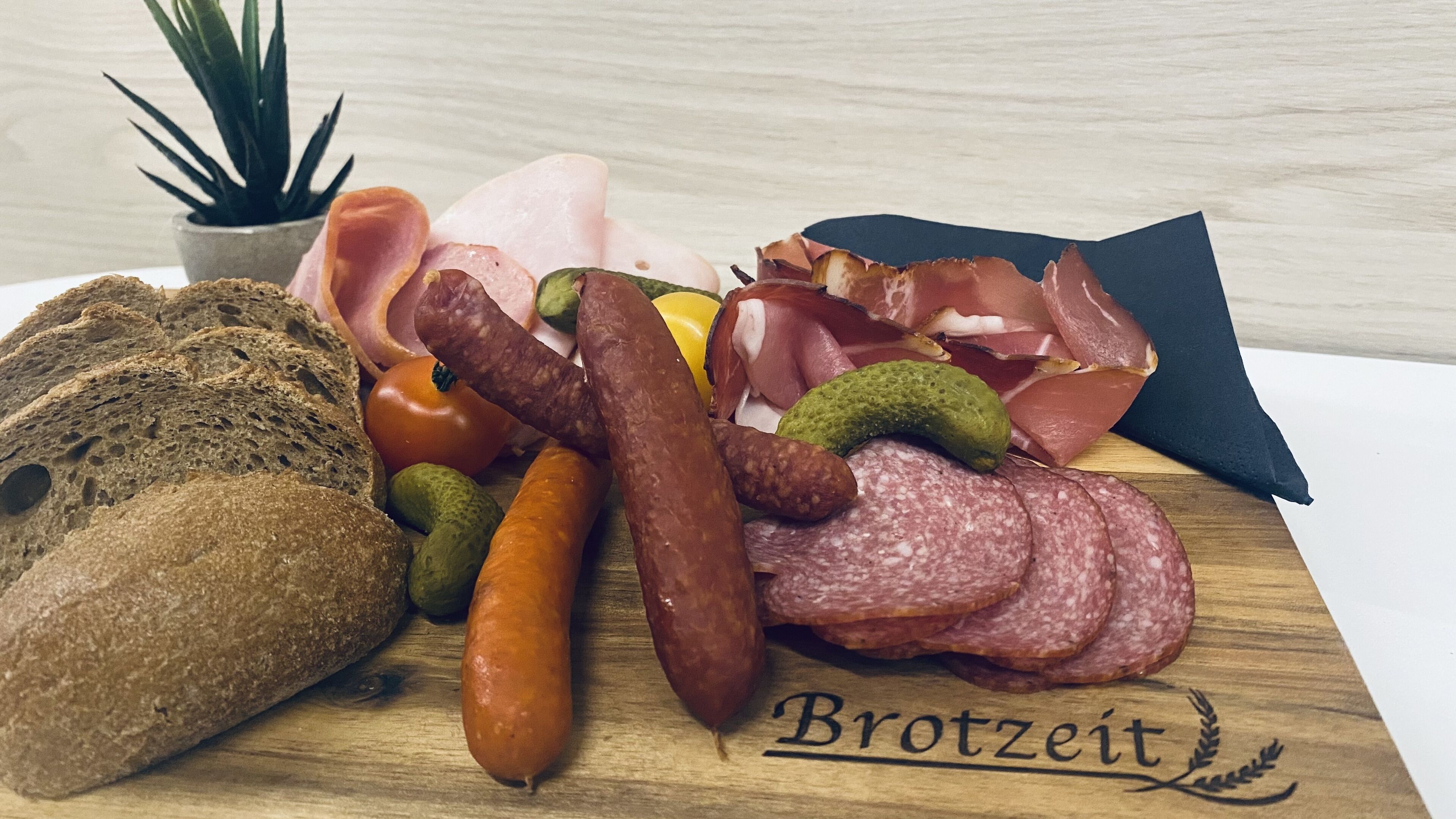 En træplanke med forskellige typer spegepølser, oste og syltede grøntsager, med teksten 'Brotzeit'.