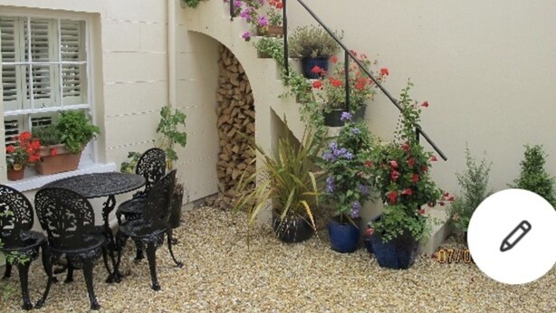 Un patio con una mesa y sillas, rodeado de plantas en macetas y una escalera con macetas colgantes de flores.