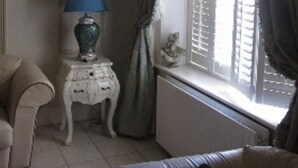 Una habitación con un sofá color crema, una mesita blanca con una lámpara azul y una ventana con persianas.