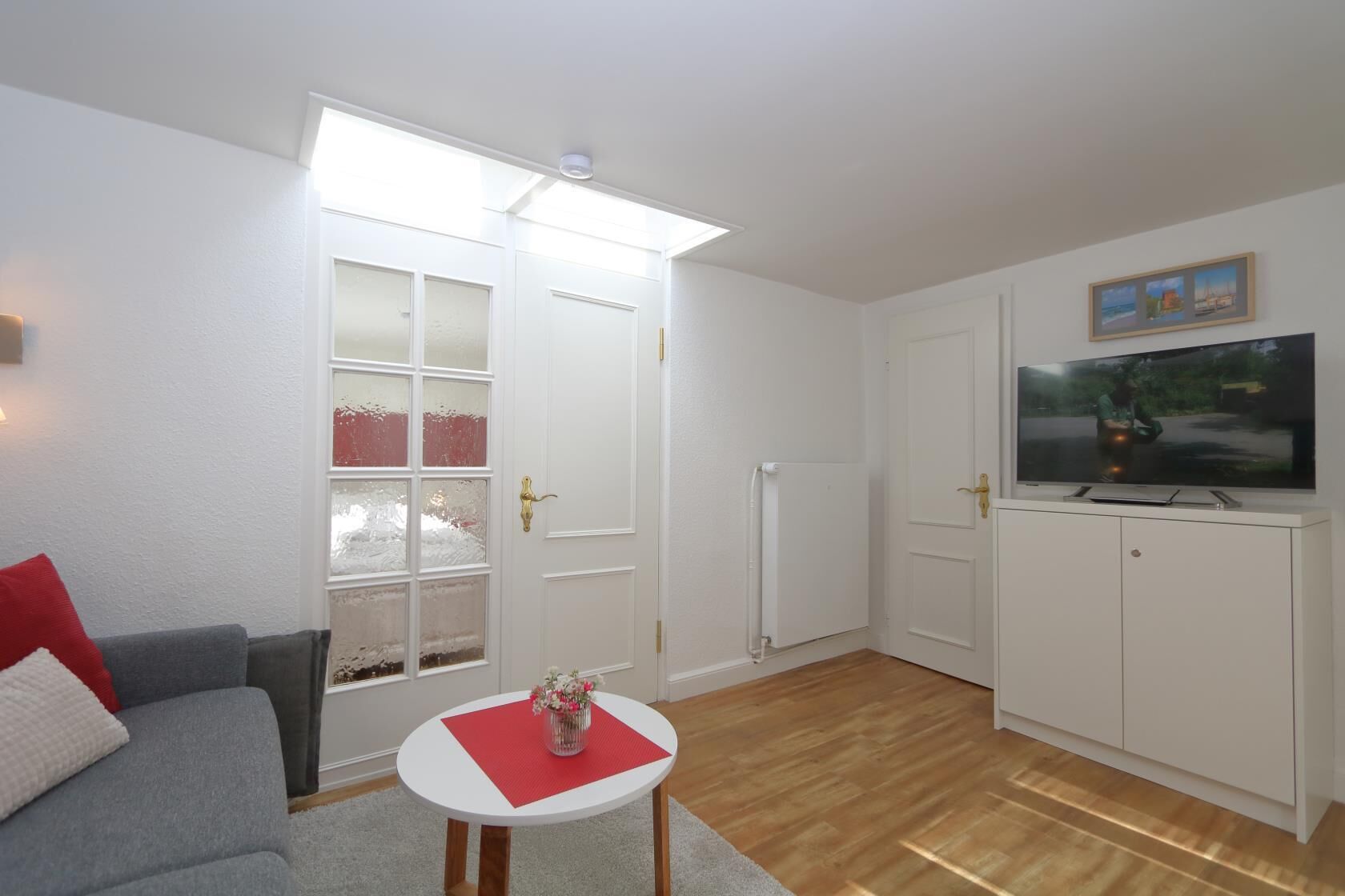 En stue med en grå sofa, et lille hvidt bord med en vase, et hvidt skab med et fjernsyn og et stort vindue med sandblæste ruder.