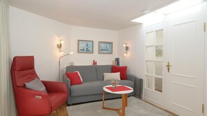 Ein Wohnzimmer mit grauem Sofa, einem roten Sessel und einem kleinen runden Tisch mit einer Vase. Zwei Wandleuchten und eine Tür mit Glasfenster sind ebenfalls vorhanden.