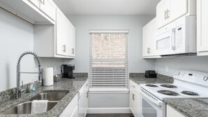 Cocina compacta con armarios blancos, encimera de granito, pileta de acero inoxidable, cafetera, estufa y ventana con persianas.