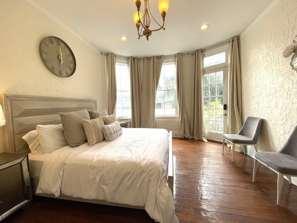 A neatly arranged bedroom with a large bed, a bedside table, a chair, a wall clock, and a window with curtains.
