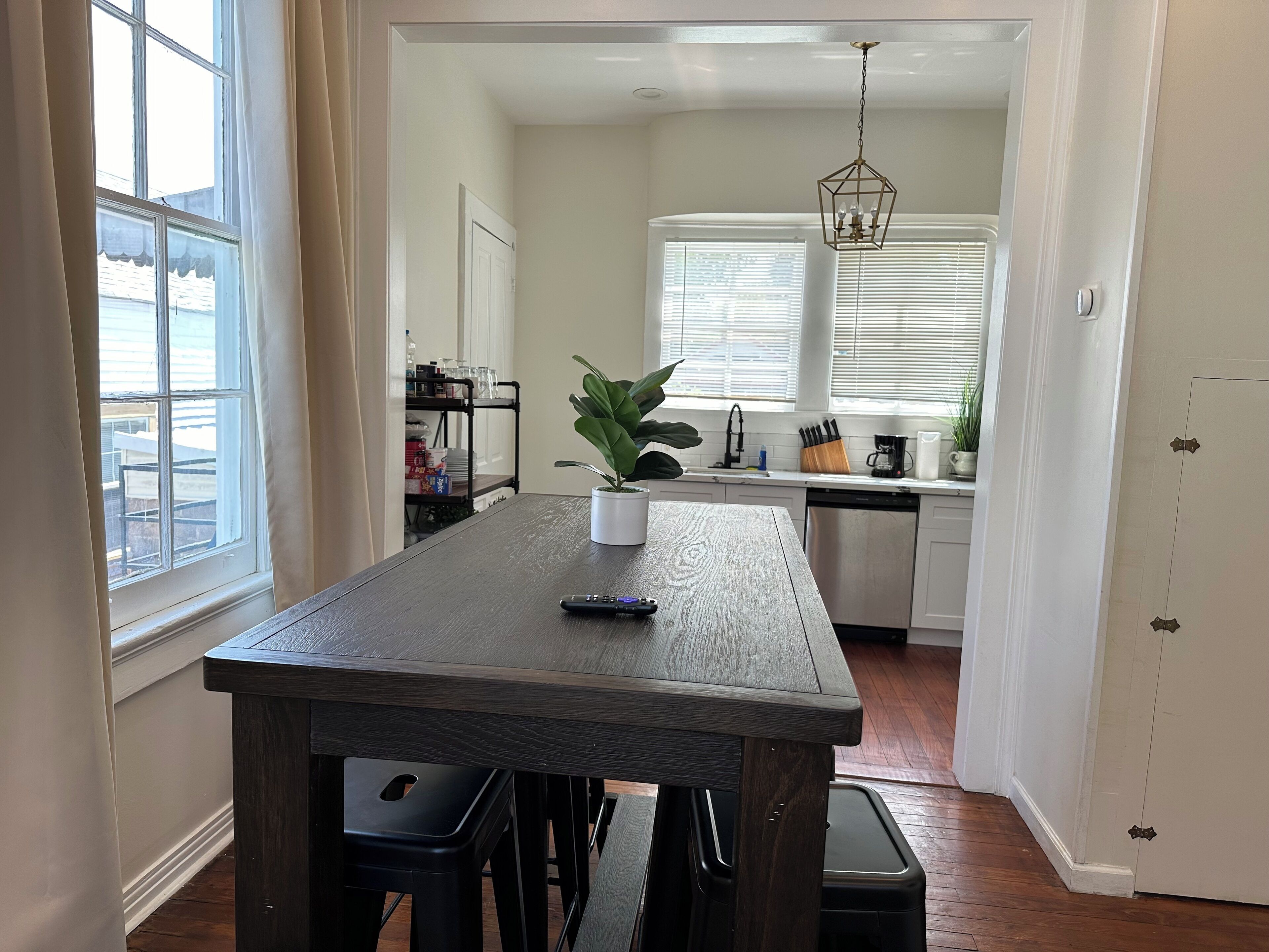A dining area with a wooden table, black stools, a potted plant, and a hanging light fixture.