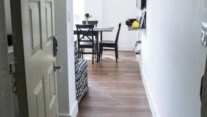 A narrow hallway with a wooden floor, a dining area with a table and chairs, and a white door on the left.