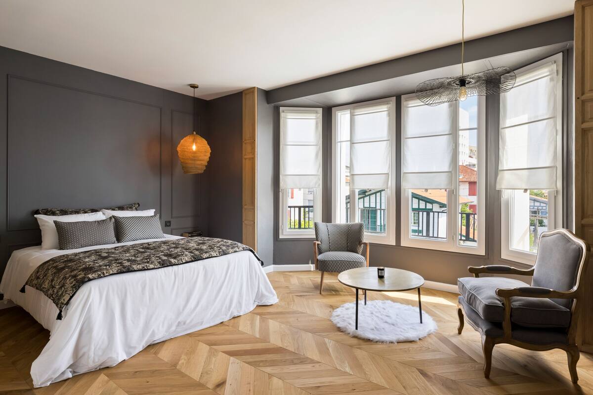 A bedroom with a large bed, a chair, a small table, and large windows with a view of buildings outside.