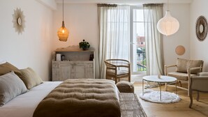 A bedroom with a large bed, a small table, two chairs, a wicker side table, and a view of the outdoors through a window. - Libertitu (Bayonne)