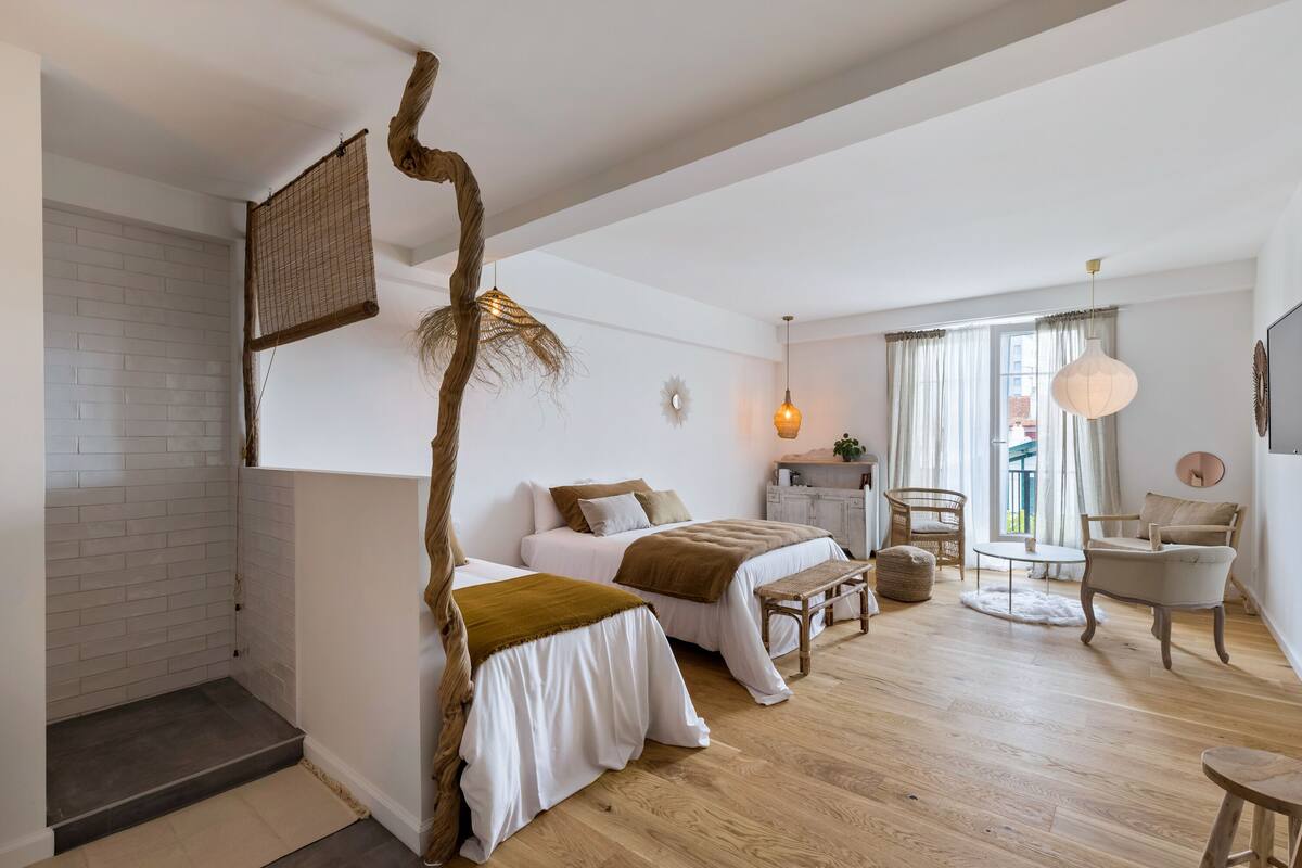 A spacious bedroom with two beds, a wooden floor, a large window with curtains, a hanging light fixture, and a rustic wall decoration.