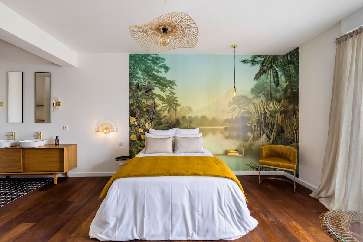 A bedroom with a large bed, a wooden dresser, a yellow chair, and a mural of a forest scene.