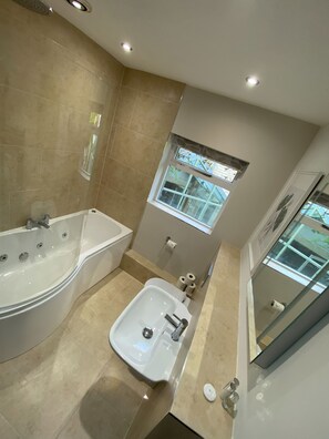 A modern bathroom with a bathtub, a sink, and a shower. There is a window with a view of greenery outside. - Church Barn (Bakewell)