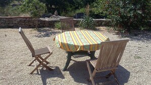A round table with a floral tablecloth, surrounded by wooden chairs, set on a gravel surface with a stone wall and greenery in the background. - Gorges de L'ardèche (Labeaume)