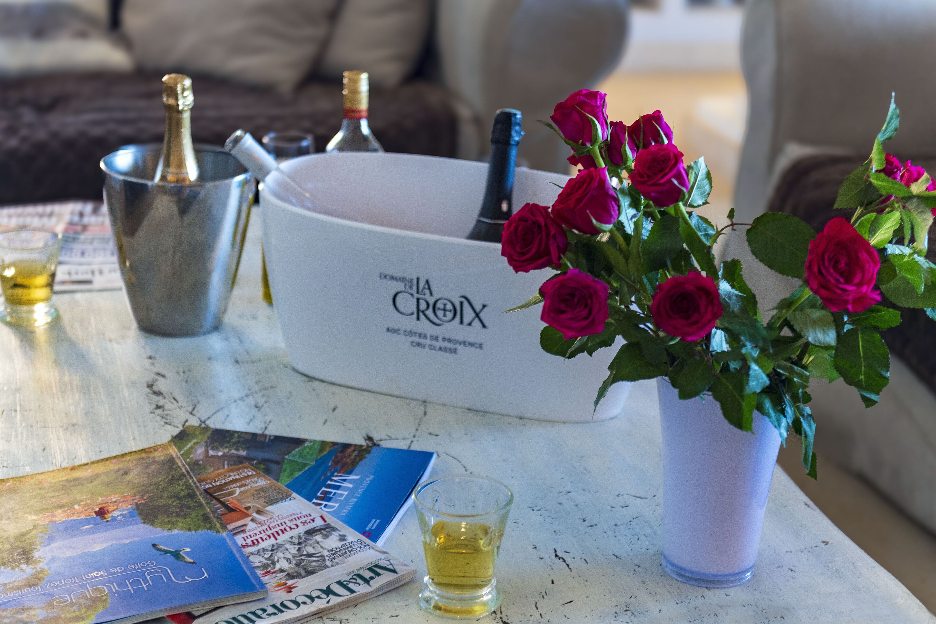 Un tavolo con una ciotola bianca con l'etichetta "La Croix", un vaso con rose rosse, un bicchierino e diverse riviste.